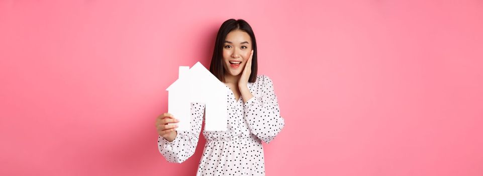 Real estate. Adult asian woman searching for home, holding house model and smiling, promo of broker company, standing over pink background.