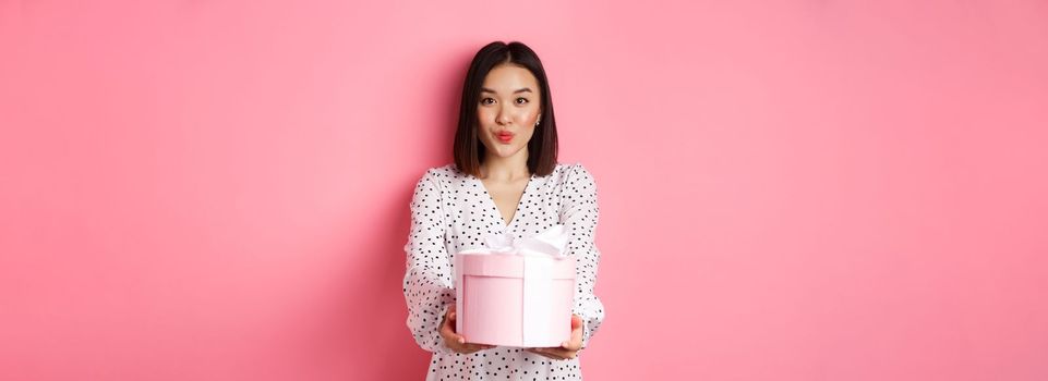 Cute asian girlfriend congratulate with valentines day, giving cute romantic gift in box, pucker lips for kiss, standing over pink background.