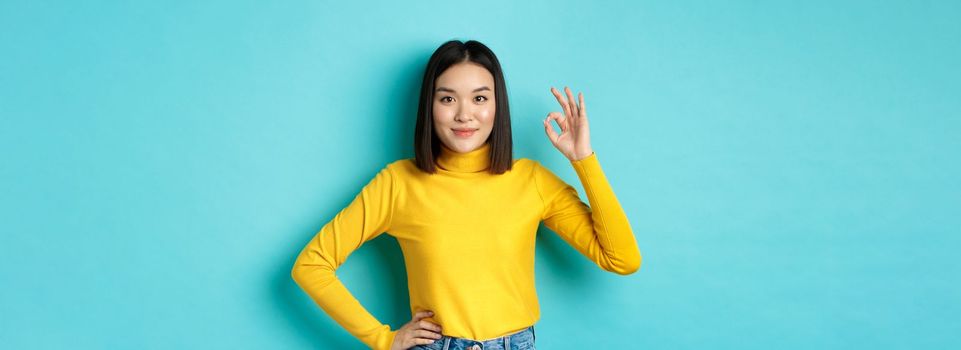 Beautiful smiling asian woman recommend product, showing Ok sign and looking satisfied, standing over blue background.