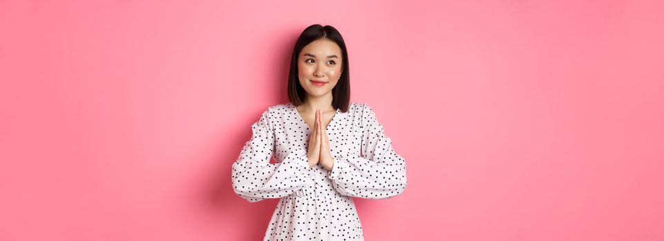 Beautiful angelic asian woman smiling, holding hands in pray and looking left at copy space with innocent cute gaze, standing over pink background.