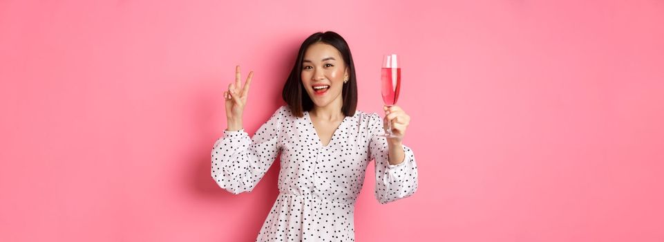 Cute asian female model drinking champagne, celebrating on party and showing peace sign, smiling happy at camera, standing over pink background.