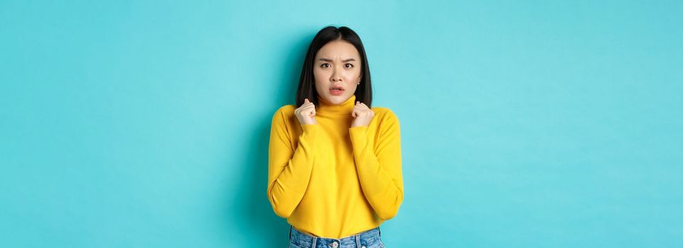 Image of worried asian woman with short dark hair, clench hands and stare at camera concerned, standing over blue background.