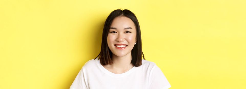 Beauty. Close up of smiling cute asian woman with perfect white smile teeth and glowing skin, standing over yellow background in t-shirt.