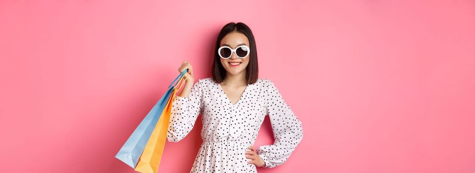 Attractive smiling asian woman holding shopping bags, wearing sunglasses and cute dress, standing against pink background.