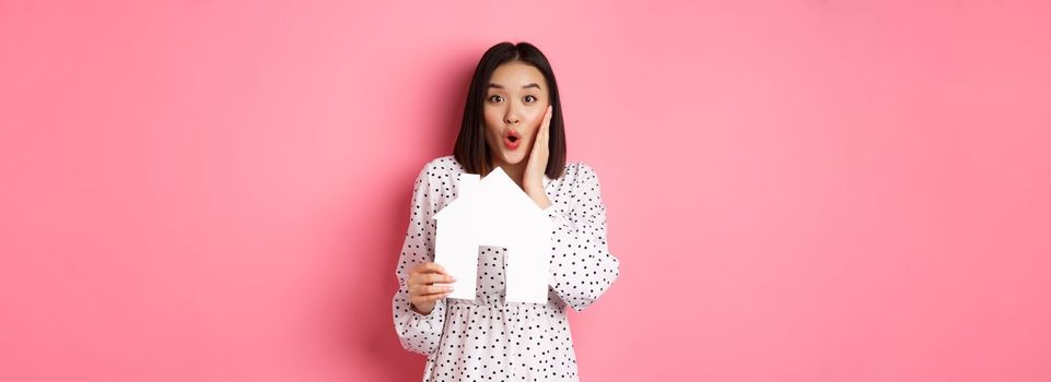 Real estate. Surprised and amazed asian woman staring at camera, saying wow and showing paper house model, standing over pink background.