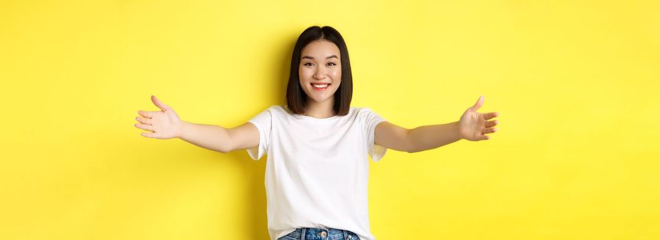 Beauty and fashion concept. Friendly asian woman spread out hands and smiling, waiting for hugs, inviting you, welcome someone and looking happy, standing over yellow background.