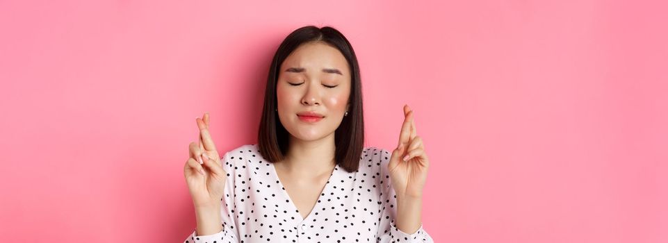 Beauty and lifestyle concept. Close-up of cute asian girl making a wish, close eyes and cross fingers hopeful, praying and standing over pink background.