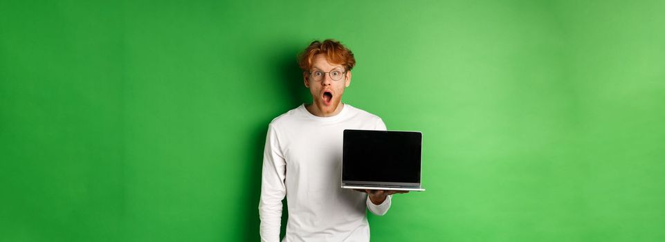 Impressed redhead guy in glasses drop jaw after seeing online promo, showing laptop screen and staring at camera with disbelief, standing over green background.