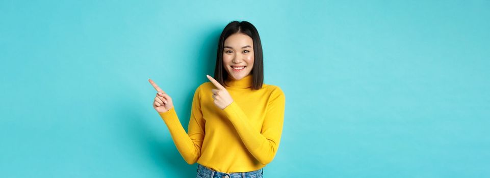 Shopping concept. Attractive asian girl in trendy sweater showing advertisement, pointing fingers right and smiling, recommend promotion deal, blue background.