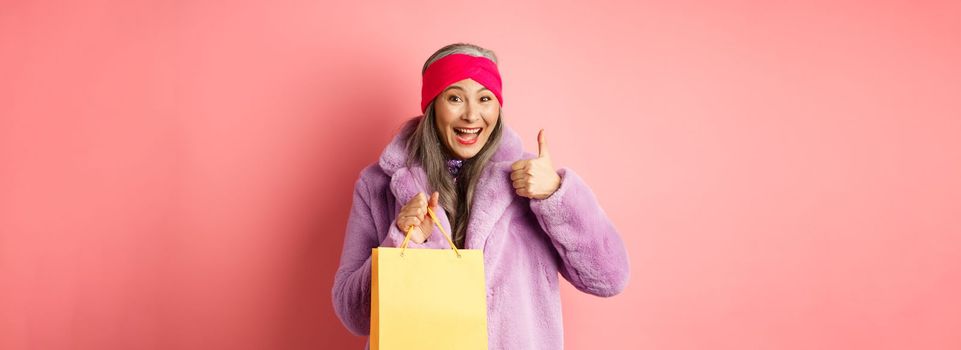 Shopping and fashion. Happy asian senior woman in stylish clothes holding paper bag from store, showing thumb-up in approval, pink background.