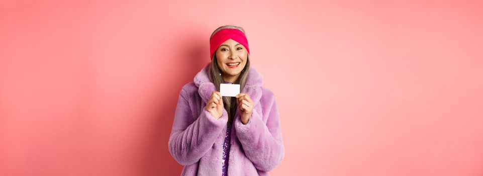 Shopping and fashion concept. Beautiful asian middle-aged woman showing plastic credit card and smiling happy in stylish clothes, pink background.