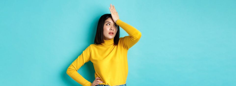 Emotions and lifestyle concept. Annoyed asian girl roll eyes and face palm, standing bothered in yellow pullover against blue background.