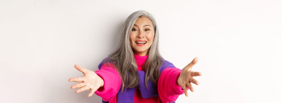 Close-up of stylish asian mature woman reaching hands forward, stretching arms for hug or hold something, smiling happy, standing over white background.