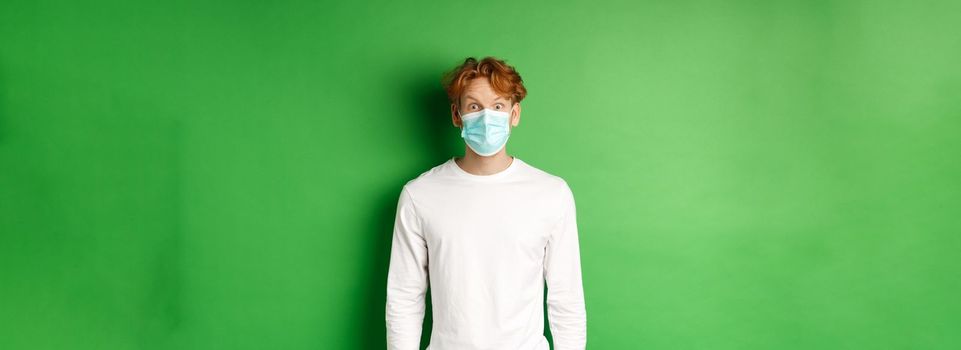 Covid-19, social distancing and lifestyle concept. Young redhead man wearing face mask during coronavirus pandemic, looking surprised at camera, green background.