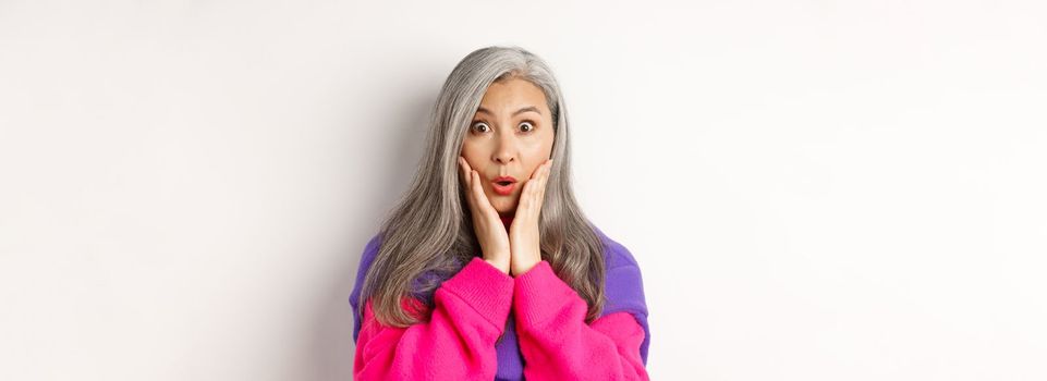 Close up of fashionable asian senior woman with grey hair looking surprised, saying wow and staring at camera amazed, standing over white background.