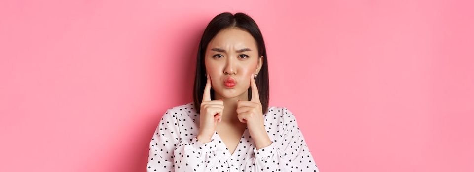 Beauty and lifestyle concept. Cute asian woman making grumpy face, poking cheeks and grimacing, standing over pink background.