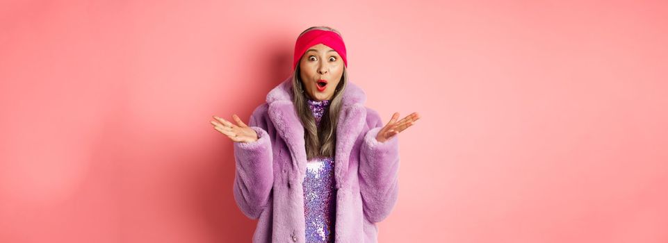 Fashion and shopping concept. Surprised elderly asian woman in stylish fake-fur coat and headband looking amazed at camera, rejoicing of good news, pink background.