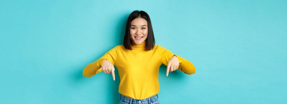 Shopping concept. Beautiful korean girl with happy smile, pointing fingers down at banner, standing against blue background.