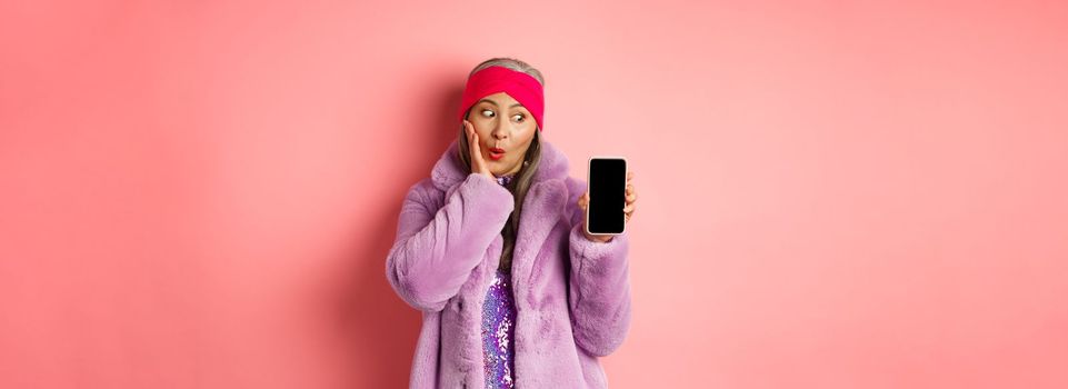 Valentines day shopping concept. Beautiful asian senior woman showing smartphone screen and looking at display excited, checking out online offer, pink background.