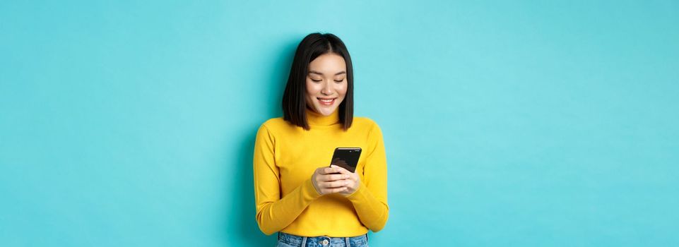 Attractive asian woman reading smartphone screen and smiling, social networking with mobile phone, standing over blue background.