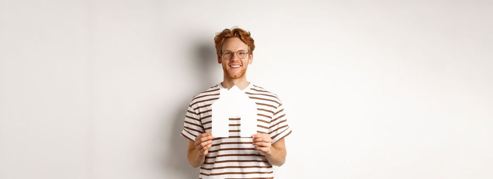 Real estate concept. Smiling redhead man in glasses holding cutout paper house and looking at camera, saving for buying property, standing over white background.