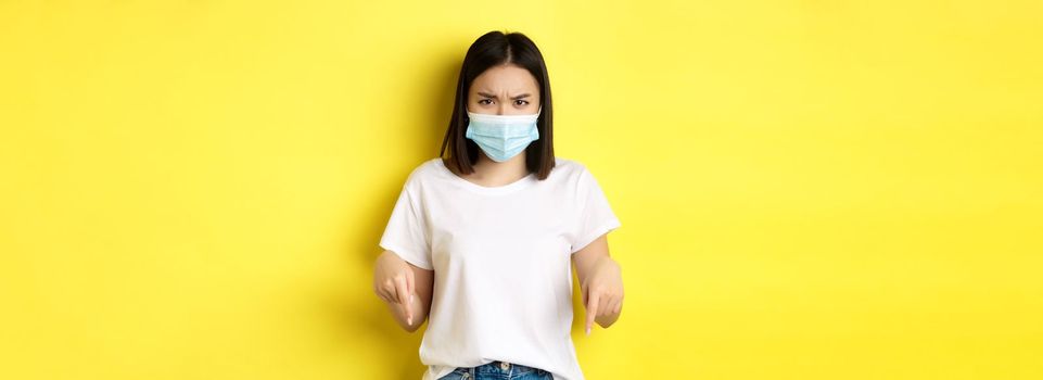 Covid-19, pandemic and social distancing concept. Disappointed asian girl in medical mask, frowning upset and pointing fingers down at logo, standing over yellow background.