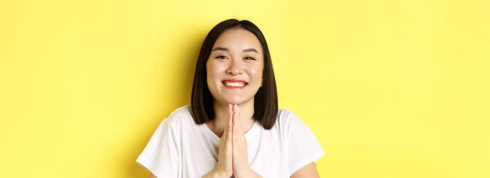 Close up of cute asian woman saying thank you and smiling, holding hands in namaste, pray gesture, standing over yellow background.