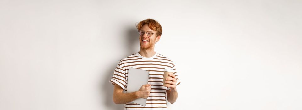 Handsome redhead male employee in glasses having break, drinking coffee and holding laptop, standing over white background.