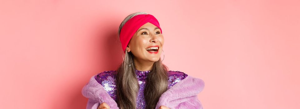 Fashion. Close-up of happy asian senior woman smiling, looking left with cheerful face, standing in purple faux fur coat, pink background.