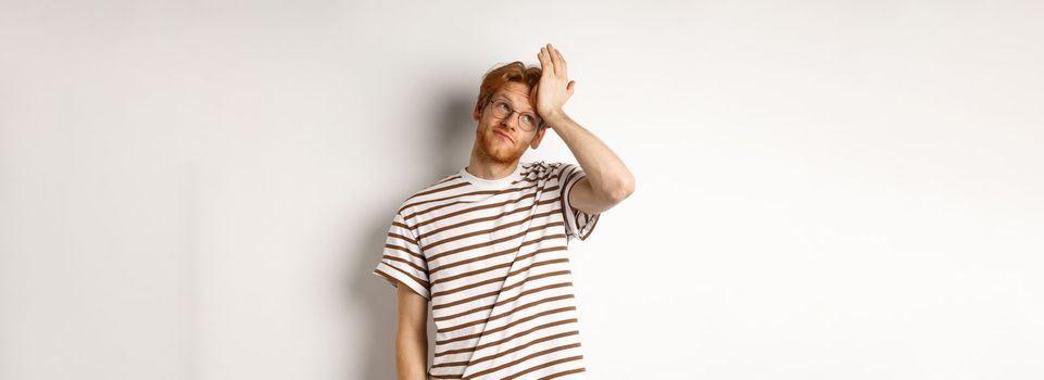 Tired young man with red hair and glasses, roll eyes and making facepalm bothered, standing over white background.
