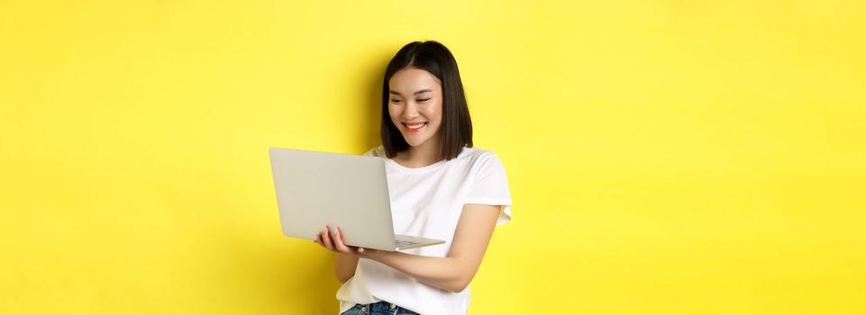 Cute asian female student working on laptop, reading screen and smiling, yellow background.