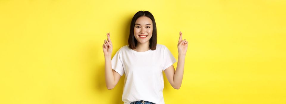 Hopeful asian girl making wish, cross fingers for good luck and praying with eyes closed, standing over yellow background.