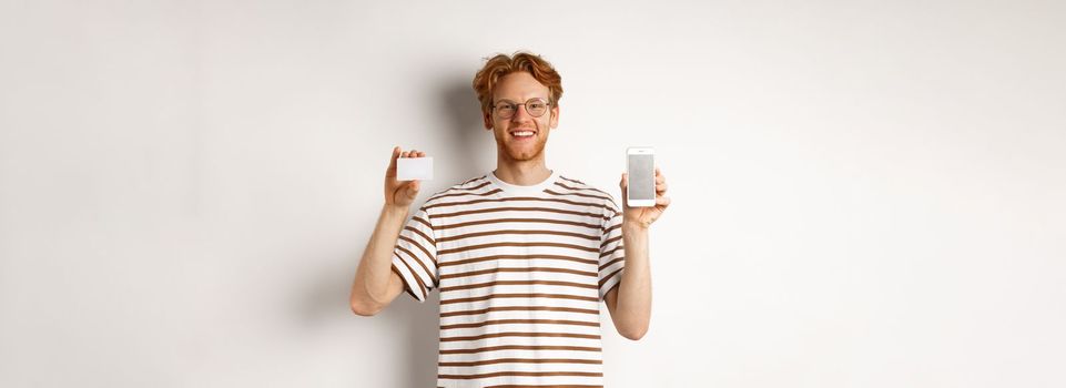 Shopping and finance concept. Young man showing blank mobile screen and plastic credit card, smiling at camera, white background.