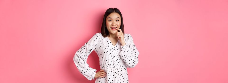 Image of beautiful young lady in dress, biting finger and smiling, staring with temptation and amazement, want buy something, standing over pink background.