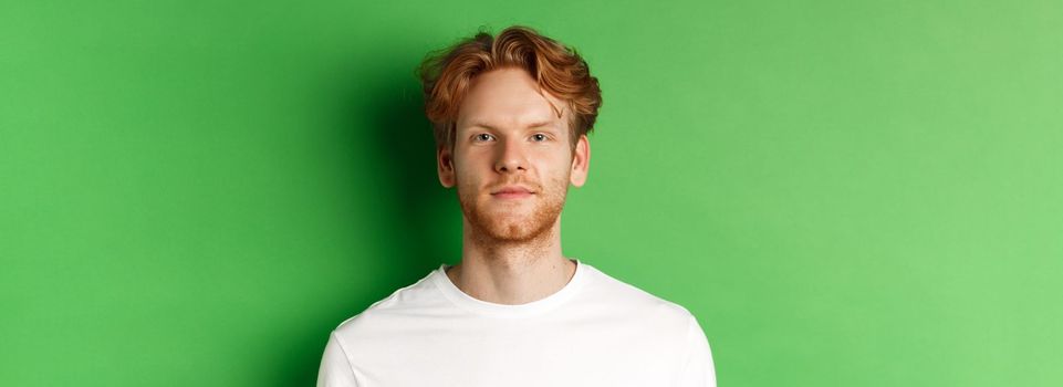 Close-up of young man with red messy hair and beard looking at camera, standing over green background.