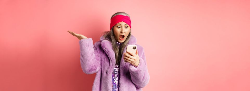 Online shopping and fashion concept. Amazed and surprised asian senior woman looking at smartphone screen, reading on mobile phone and gasping, pink background.
