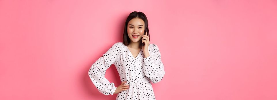 Beautiful asian woman having mobile conversation, making phone call and smiling, standing over pink background.