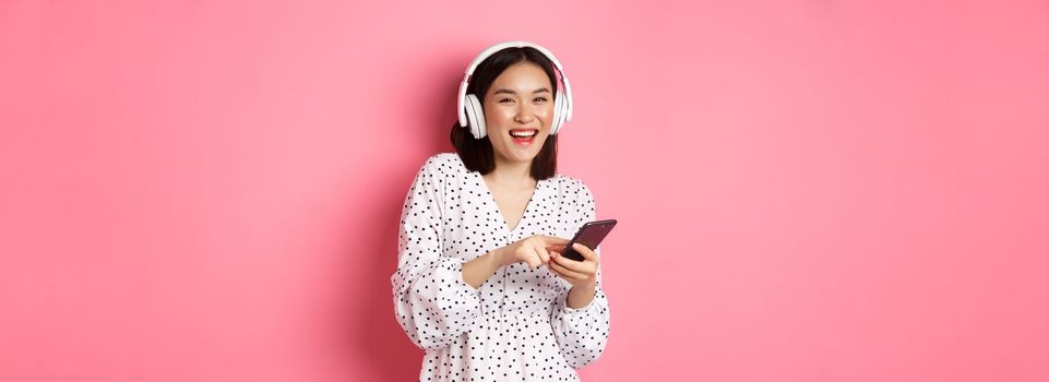 Beautiful asian woman listening music in headphones, using mobile phone, smiling happy at camera, standing over pink background.