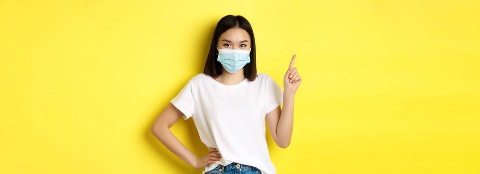 Covid, health care and pandemic concept. Asian female model in medical mask and white t-shirt pointing finger at upper left corner logo, showing promotion, yellow background.