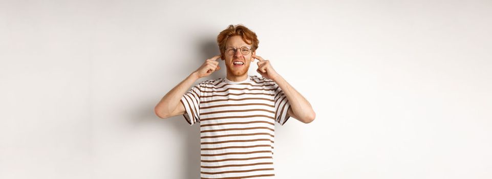 Annoyed redhead guy in glasses shutting ears and complaining loud noise, angry at noisy neighbours, standing over white background.