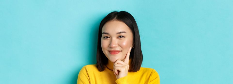 Beauty and makeup concept. Close up of thoughtful asian woman looking pleased at camera and smile, having an idea, standing over blue background.