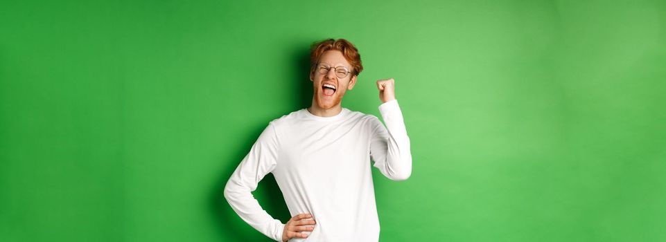 Young cheerful guy scream of joy, winning prize and celebrating, making fist pump gesture and smiling happy, standing over green background.
