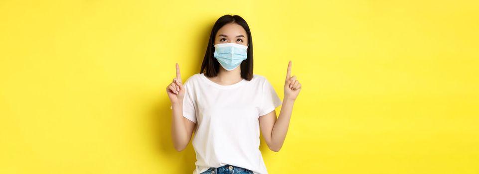 Covid-19, pandemic and social distancing concept. Young asian woman in white t-shirt and medical mask from coronavirus, looking and pointing fingers up, showing special offer.