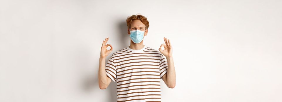 Covid-19, pandemic and social distancing concept. Young man with red hair wearing medical mask to prevent catching coronavirus, showing okay signs in approval, white background.