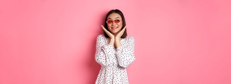 Tourism and lifestyle concept. Beautiful asian woman showing her clean cute face, wearing sunglasses, smiling and looking aside with admiration, standing over pink background.