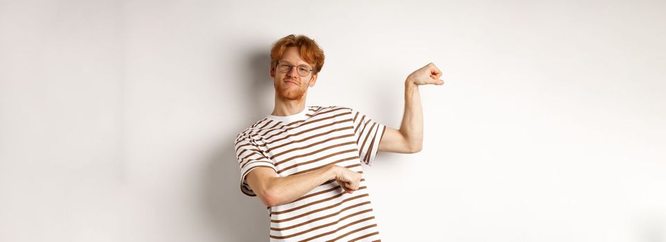 Image of confident and strong redhead man flexing biceps, showing muscles after gym, standing over white background.