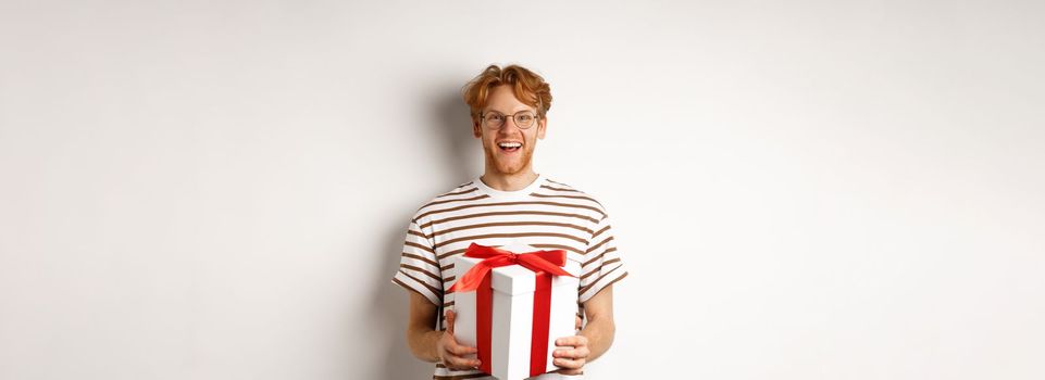 Valentines day and holidays concept. Cheerful young man holding gift box and smiling grateful, receiving presents, standing over white background.