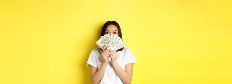 Cute asian woman hiding face behind money, peeking at camera satisfied, earn cash, standing over yellow background.