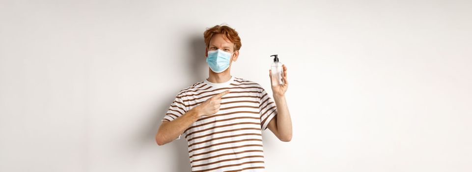 Covid-19, health and lifestyle concept. Cheerful redhead man in face mask pointing finger at hand sanitizer, recommending antiseptic, white background.