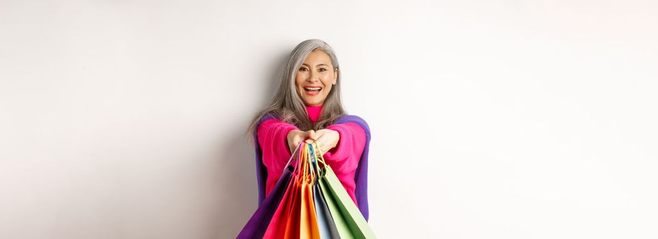 Fashionable asian senior woman going on shopping, extending hands with paper bags, smiling satisfied at camera, standing over white background.